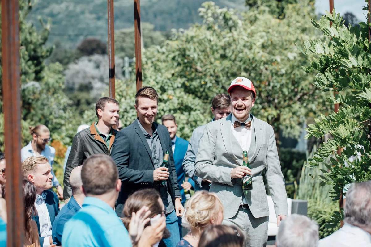 Lachender Bräutigam mit Freunden und Bierflaschen im Garten, vorne sitzende Gäste
