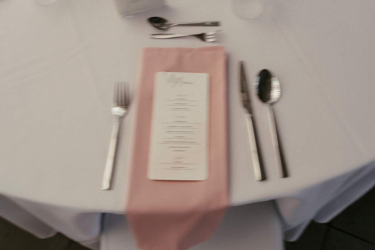 Gedeck mit rosa Serviette, Menükarte und Besteck auf weißer Tischdecke im Gewächshaus